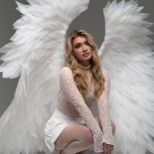 May include: A woman wearing a white lace dress and white tights poses with large white angel wings. She is wearing a pearl necklace and has long blonde hair.