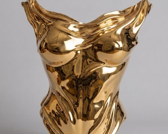 Op de afbeelding: Een gepolijst goudkleurig beeldhouwwerk in de vorm van een menselijke torso. Het oppervlak is sterk reflecterend en heeft vloeiende, golvende contouren die het uiterlijk van gedrapeerde stof of gesmolten metaal nabootsen.