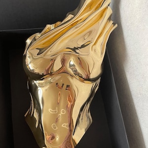 Op de afbeelding: Een gepolijst goudkleurig beeldhouwwerk van een menselijke torso met een reflecterend, metalen oppervlak en vloeiende, gedrapeerde contouren. Het object heeft een gladde, hoogglanzende afwerking die licht vangt en vervormde reflecties cre&euml;ert.