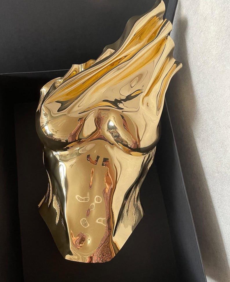 Op de afbeelding: Een gepolijst goudkleurig beeldhouwwerk van een menselijke torso met een reflecterend, metalen oppervlak en vloeiende, gedrapeerde contouren. Het object heeft een gladde, hoogglanzende afwerking die licht vangt en vervormde reflecties cre&euml;ert.