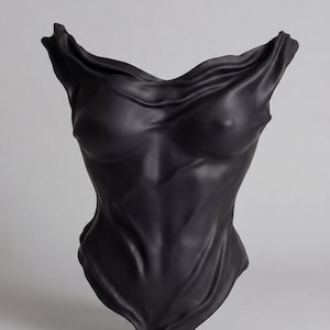 Op de afbeelding: Een matzwart keramieken sculptuur in de vorm van een menselijke torso. Het oppervlak vertoont diepe, vloeiende plooien en vouwen die het uiterlijk van gedrapeerde stof nabootsen, wat een getextureerde, gevormde figuur met een wijde halslijn cre&euml;ert.