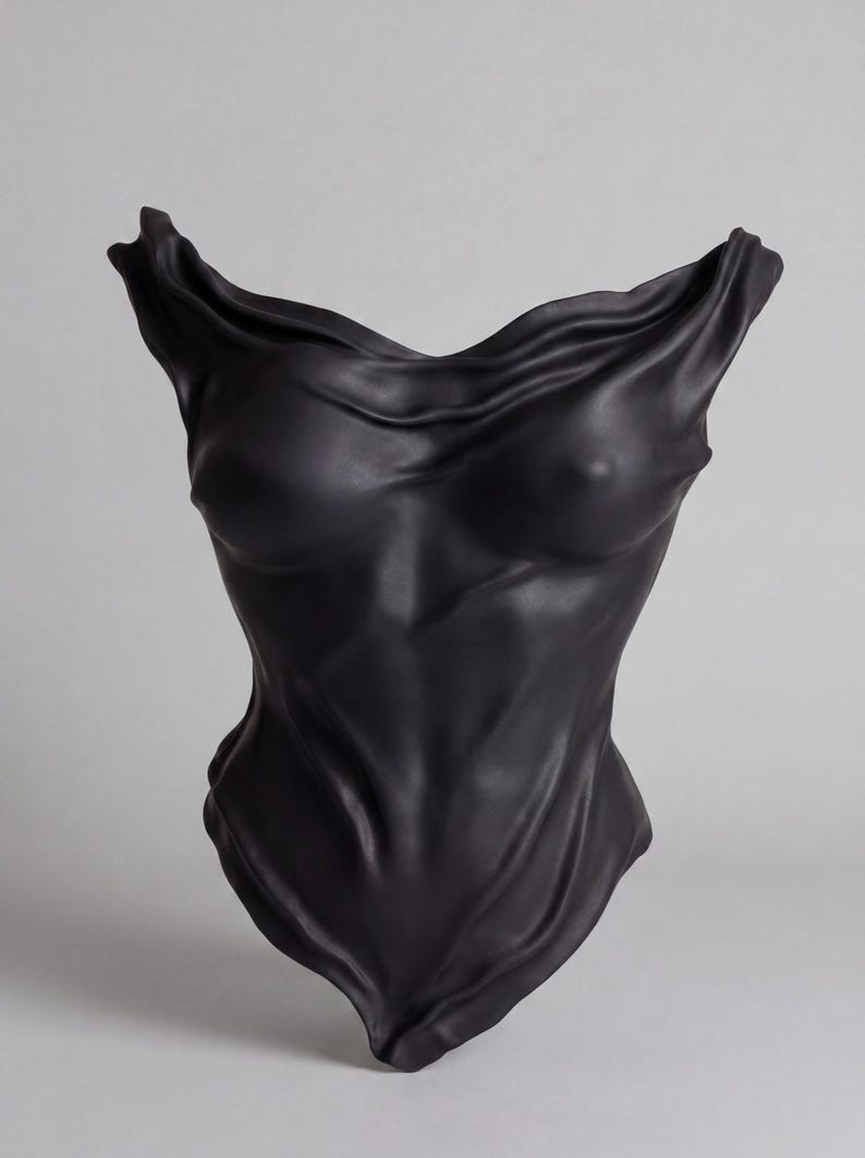 Op de afbeelding: Een matzwart keramieken sculptuur in de vorm van een menselijke torso. Het oppervlak vertoont diepe, vloeiende plooien en vouwen die het uiterlijk van gedrapeerde stof nabootsen, wat een getextureerde, gevormde figuur met een wijde halslijn cre&euml;ert.