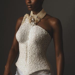 May include: A textured white one-shoulder corset with a draped skirt. The model wears a pearl and jeweled choker necklace. The overall aesthetic is elegant and high fashion, suitable for special occasions.