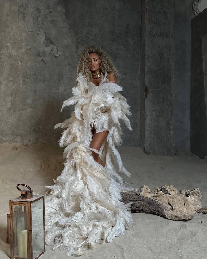 K&ouml;nnte beinhalten: Eine Frau in einem wei&szlig;en Federkleid und einer goldenen Halskette. Sie steht in einem sandigen Bereich mit einem Holzstamm und einer Laterne.
