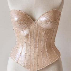 May include: A strapless, peach-colored corset with vertical ribbed detailing and subtle bead embellishments. The bodice is structured with a sweetheart neckline and a fitted silhouette, showcasing a delicate and elegant design.
