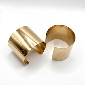 Puede incluir: Dos pulseras de puño de metal dorado. Las pulseras son abiertas y tienen una superficie lisa y reflectante. Los puños son anchos y cilíndricos, diseñados para ser usados en la muñeca. Las joyas se exhiben sobre una superficie blanca.