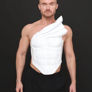 Könnte beinhalten: Ein weißes, geformtes Bruststück mit asymmetrischem Design. Das Kleidungsstück wird über dem Oberkörper getragen, mit einem drapierten Stoffelement über einer Schulter. Die Person trägt eine schwarze Hose und ein silbernes Armband. Der Hintergrund ist schwarz.