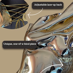 Op de afbeelding: Een metallic zilveren sculpturaal kledingstuk met een reflecterende, vloeistofachtige textuur. Een inzetstuk toont een zwart stoffen gedeelte met een verstelbare vetersluiting aan de achterkant en goudkleurige koorden. Tekst: Important to know.