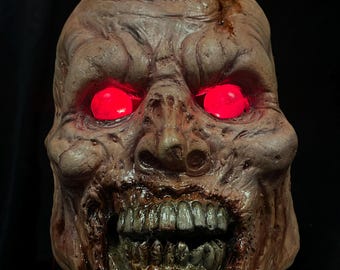 May include: A gruesome zombie mask with glowing red eyes. The mask is a mix of brown and tan tones, with exposed teeth and a decaying appearance. The eyes glow brightly, adding to the frightening effect. Perfect for Halloween or horror events.