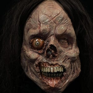May include: A detailed, gruesome mask with a decaying, flesh-toned face. The mask features a single, open eye, exposed teeth, and dark hair. The face has visible cracks and a realistic, unsettling appearance, perfect for Halloween.