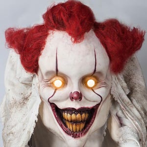Op de afbeelding: Een griezelig clownmasker met felrood haar, een wit gezicht en felgele ogen. De clown heeft een sinistere grijns, donkerrode lippen en gele tanden. Het masker is onderdeel van een Halloween-kostuum of rekwisiet.