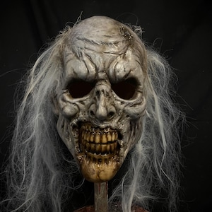 Op de afbeelding: Een gedetailleerd, verweerd zombiemasker met een grijs, gerimpeld gezicht, verzonken oogkassen en een vervallen, blootliggende kaak. Het masker heeft lang, wit, ijl haar en is gemonteerd op een metalen standaard.