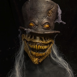 May include: A full-head mask featuring a dark brown, weathered top hat with frayed fabric accents. The face has a wide, toothy grin with yellowed teeth, glowing orange eyes, and long, thin, wispy grey hair strands hanging from the sides.