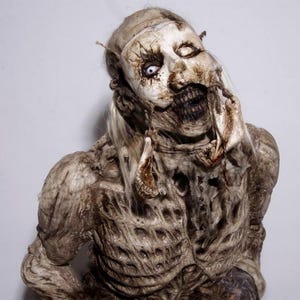 Può includere: Primo piano dettagliato di una figura di zombie con pelle in decomposizione e denti scoperti. La figura ha un incarnato pallido, con un occhio aperto e l'altro chiuso. Il costume ha un aspetto lacerato, con fili di capelli e tessuto che pendono dalla testa e dalle spalle.
