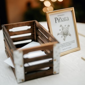 May include: A wooden crate with a white card inside and a gold frame with a sign that says "Anniversary Pi&ntilde;ata" with a drawing of a pinata in the shape of a star.