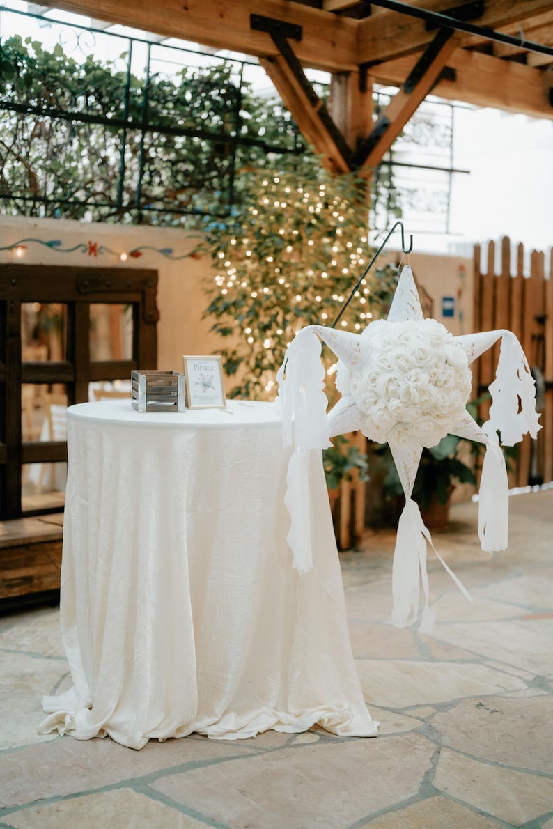 May include: A white table with a white tablecloth is set up in a room with a white pinata hanging from the ceiling. The pinata is shaped like a star and is decorated with white flowers. A small sign on the table reads "Drinks".