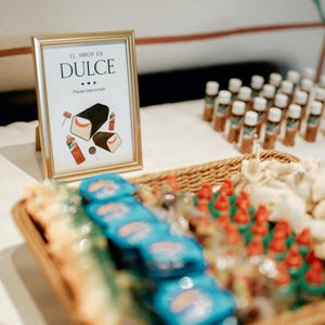 May include: A gold-framed sign with the words "EL AMOR ES DULCE" and illustrations of treats. A woven basket holds blue-wrapped sweets and red-topped items. Small bottles with white caps are arranged in rows.