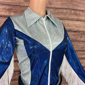 Puede incluir: Una camisa de manga larga con acabado azul met&aacute;lico brillante en las mangas y el torso, contrastada por un canes&uacute; y cuello plateados iridiscentes y texturizados. La prenda incluye una cremallera frontal blanca y flecos blancos largos.