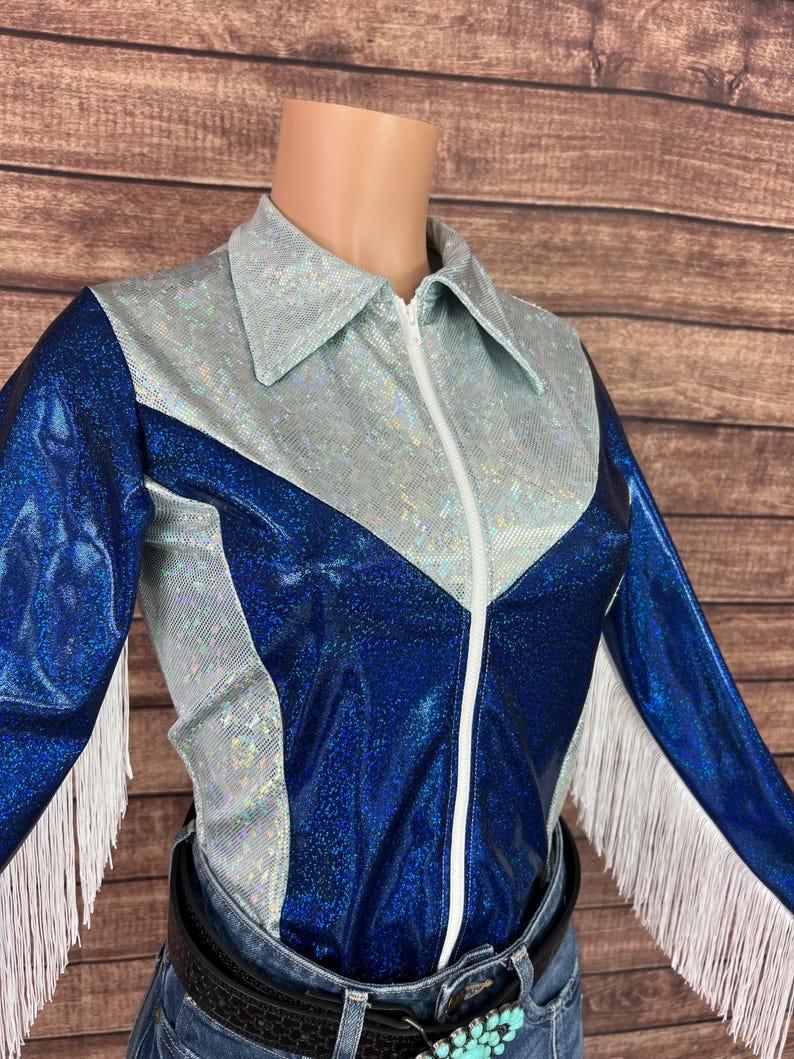 Puede incluir: Una camisa de manga larga con acabado azul met&aacute;lico brillante en las mangas y el torso, contrastada por un canes&uacute; y cuello plateados iridiscentes y texturizados. La prenda incluye una cremallera frontal blanca y flecos blancos largos.