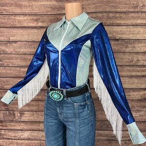 Puede incluir: Una camisa de manga larga con un dise&ntilde;o de bloques de color azul met&aacute;lico y plata iridiscente con cremallera frontal, flecos blancos a lo largo de las mangas y un cintur&oacute;n negro con una gran hebilla ovalada de color turquesa sobre vaqueros azules.
