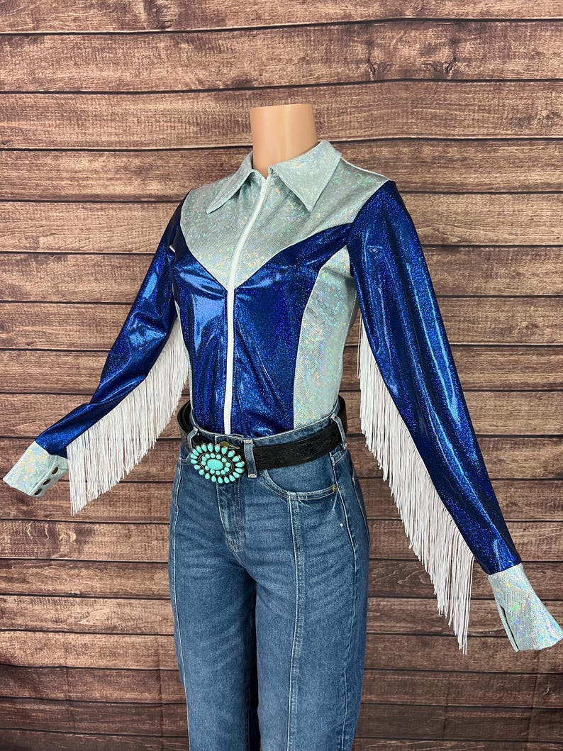 Puede incluir: Una camisa de manga larga con un dise&ntilde;o de bloques de color azul met&aacute;lico y plata iridiscente con cremallera frontal, flecos blancos a lo largo de las mangas y un cintur&oacute;n negro con una gran hebilla ovalada de color turquesa sobre vaqueros azules.