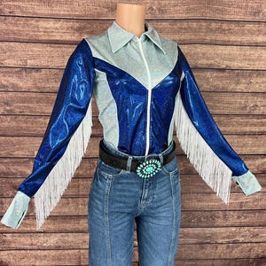 Puede incluir: Una camisa de manga larga con un dise&ntilde;o de bloques de color azul met&aacute;lico y plata iridiscente, cremallera frontal, cuello en punta y flecos blancos a lo largo de las mangas, combinada con pantalones vaqueros azules y un cintur&oacute;n negro con hebilla turquesa.