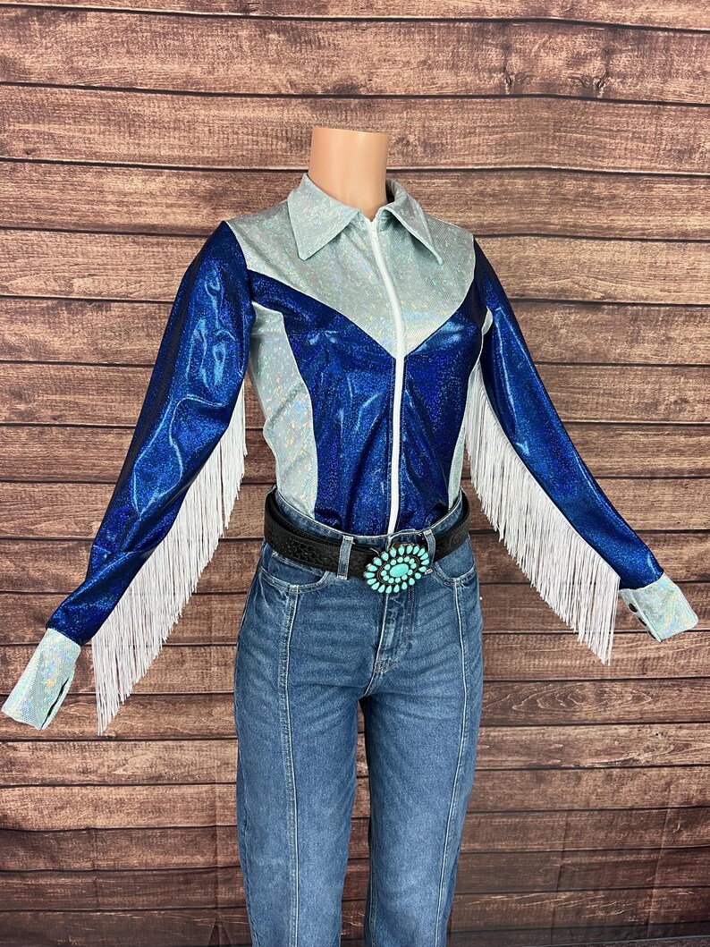 Puede incluir: Una camisa de manga larga con un dise&ntilde;o de bloques de color azul met&aacute;lico y plata iridiscente, cremallera frontal, cuello en punta y flecos blancos a lo largo de las mangas, combinada con pantalones vaqueros azules y un cintur&oacute;n negro con hebilla turquesa.