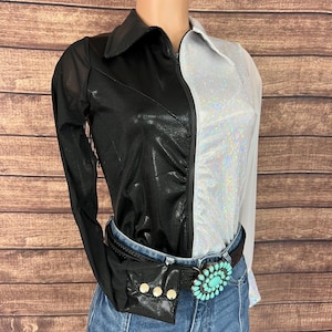 May include: A two-toned top with a black faux leather look on the left and a silver, sparkly fabric on the right. The top has a zipper closure and is paired with a black belt with a turquoise-colored buckle and a small black fanny pack. The model is wearing blue jeans.