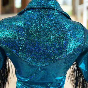 Puede incluir: Una camisa azul turquesa con un acabado brillante e iridiscente. La camisa tiene un yugo de estilo occidental y ribetes de flecos negros en las mangas.