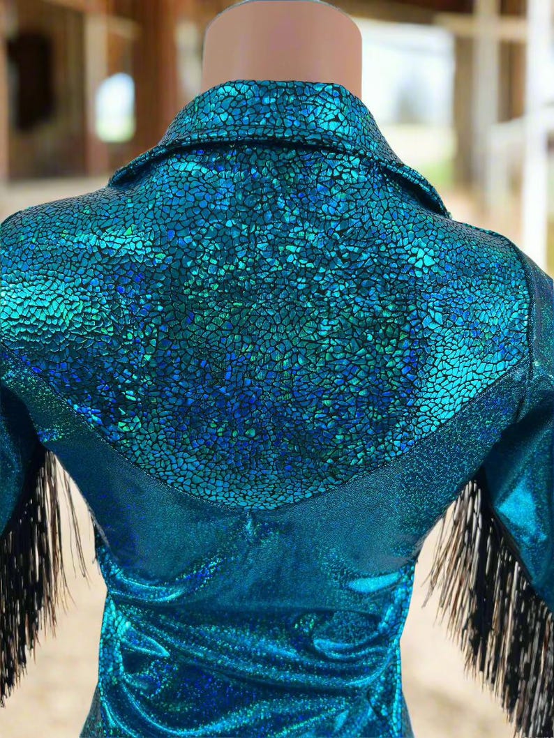 Puede incluir: Una camisa azul turquesa con un acabado brillante e iridiscente. La camisa tiene un yugo de estilo occidental y ribetes de flecos negros en las mangas.