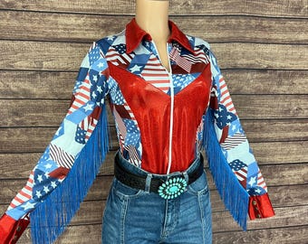 May include: A long-sleeve shirt featuring a red, white, and blue American flag pattern with solid red panels on the chest, blue fringe along the sleeves, and a white zipper. The shirt is paired with blue denim jeans and a black belt with a turquoise buckle.