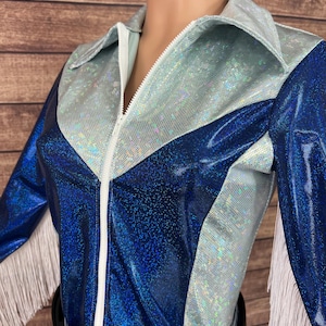 Puede incluir: Una camisa de manga larga con un dise&ntilde;o de tela hologr&aacute;fica en azul met&aacute;lico y plata iridiscente. La prenda incluye un cierre de cremallera frontal blanco, un cuello puntiagudo y detalles de flecos blancos en los bordes de las mangas.