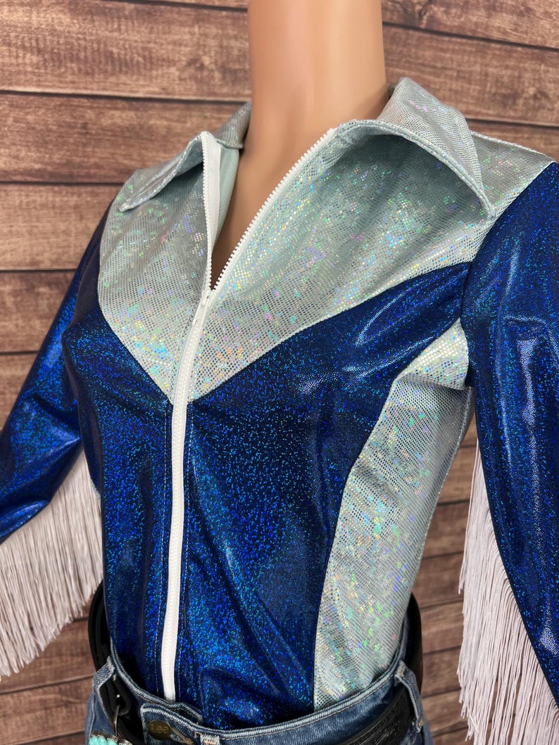 Puede incluir: Una camisa de manga larga con un dise&ntilde;o de tela hologr&aacute;fica en azul met&aacute;lico y plata iridiscente. La prenda incluye un cierre de cremallera frontal blanco, un cuello puntiagudo y detalles de flecos blancos en los bordes de las mangas.