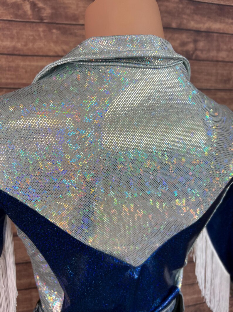 Puede incluir: Una camisa de manga larga con un panel superior texturizado hologr&aacute;fico plateado con cuello y una secci&oacute;n inferior met&aacute;lica azul oscuro. La prenda incluye detalles de flecos blancos a lo largo de las mangas y una costura en forma de V.