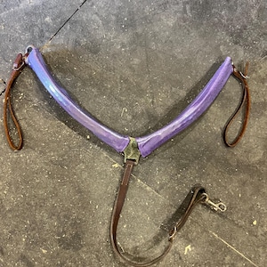 May include: A purple horse collar with brown leather straps and a metal clasp. The collar is V-shaped and has a metallic sheen. The leather straps are attached to the collar and are designed to be connected to a horse's harness.
