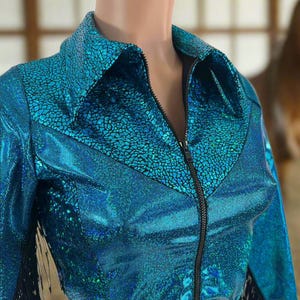 Puede incluir: Una chaqueta azul turquesa con cremallera negra y flecos negros. La chaqueta tiene un acabado brillante e iridiscente.