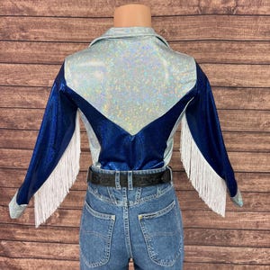 Puede incluir: Una camisa de manga larga con una secci&oacute;n superior hologr&aacute;fica plateada y mangas y panel trasero en azul met&aacute;lico. La prenda incluye flecos blancos a lo largo de los brazos y se combina con pantalones vaqueros azules y un cintur&oacute;n negro.