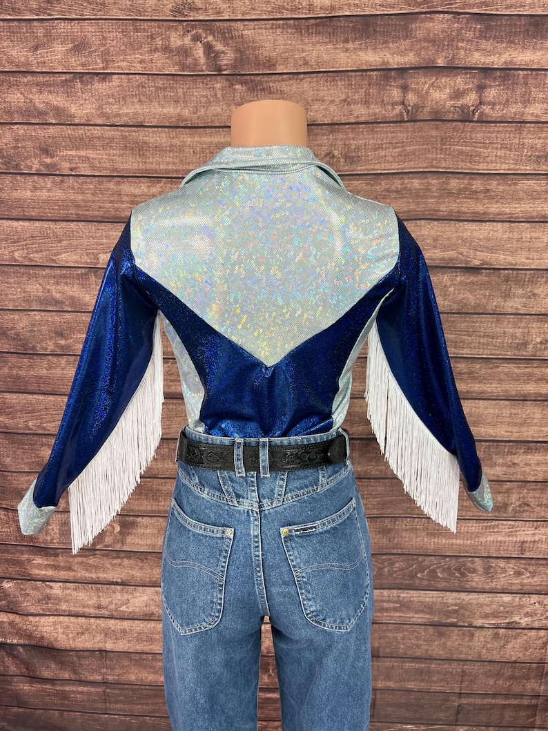Puede incluir: Una camisa de manga larga con una secci&oacute;n superior hologr&aacute;fica plateada y mangas y panel trasero en azul met&aacute;lico. La prenda incluye flecos blancos a lo largo de los brazos y se combina con pantalones vaqueros azules y un cintur&oacute;n negro.