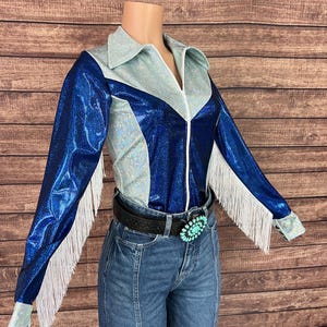 Puede incluir: Una camisa de manga larga con un dise&ntilde;o de bloques de color en paneles azul met&aacute;lico y azul claro iridiscente. La camisa incluye un cuello en punta, una cremallera frontal y detalles de flecos blancos en las mangas, combinada con jeans azules.