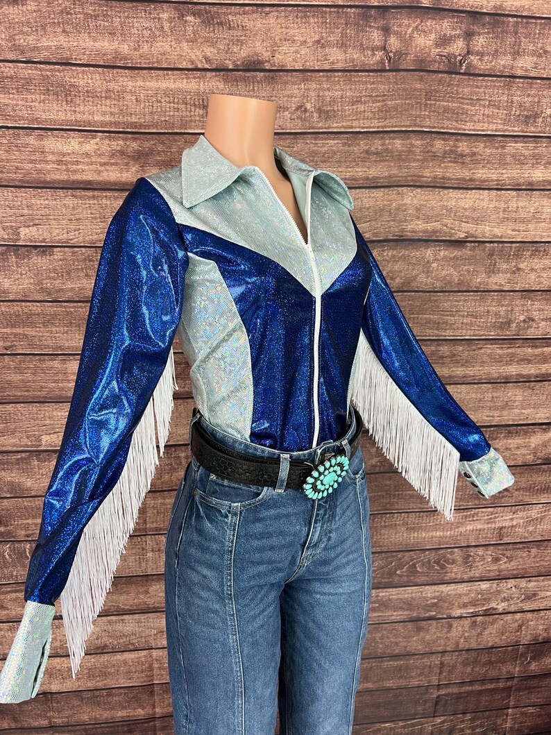 Puede incluir: Una camisa de manga larga con un dise&ntilde;o de bloques de color en paneles azul met&aacute;lico y azul claro iridiscente. La camisa incluye un cuello en punta, una cremallera frontal y detalles de flecos blancos en las mangas, combinada con jeans azules.