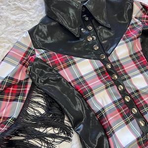 May include: A black and red plaid shirt with a black leather collar and sleeves. The shirt has silver buttons and black fringe along the sleeves. The plaid pattern includes red, white, and green lines.