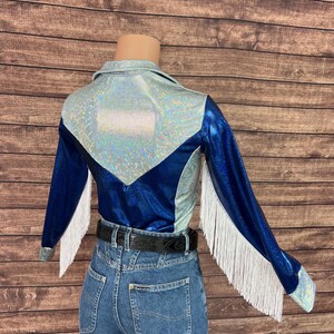 Puede incluir: Una camisa de manga larga con una secci&oacute;n superior hologr&aacute;fica plateada y una secci&oacute;n inferior azul met&aacute;lico profundo con una costura en forma de V. Las mangas est&aacute;n decoradas con largos flecos blancos, combinada con pantalones vaqueros azules y un cintur&oacute;n negro.