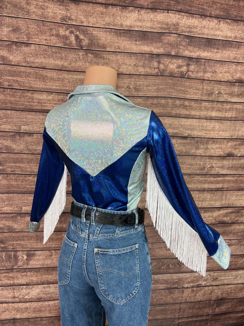 Puede incluir: Una camisa de manga larga con una secci&oacute;n superior hologr&aacute;fica plateada y una secci&oacute;n inferior azul met&aacute;lico profundo con una costura en forma de V. Las mangas est&aacute;n decoradas con largos flecos blancos, combinada con pantalones vaqueros azules y un cintur&oacute;n negro.