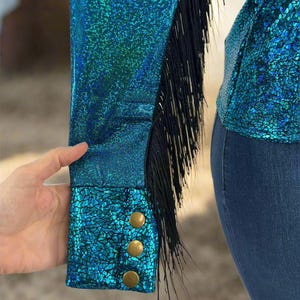 Puede incluir: Una camisa azul turquesa con ribetes de flecos negros y botones dorados en los pu&ntilde;os. La camisa tiene un acabado brillante e iridiscente.