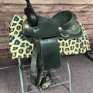 May include: A black leather Western saddle with a horn, stirrups, and a seat. The saddle is resting on a leopard print pad. The saddle is supported by a metal stand.