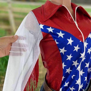 May include: A red, white, and blue zip-up top with a star pattern and white mesh sleeves. The top has a collar and a white zipper. Red fringe adorns the sleeve. The top is worn with a brown belt and blue jeans.