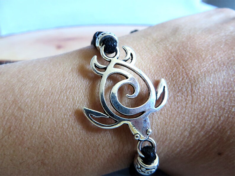 May include: A black cord bracelet with a silver turtle charm. The turtle is a stylized design with a swirl in the center.