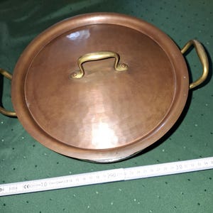 Puede incluir: Una olla de cobre con tapa y dos asas. La olla tiene un diámetro de unos 30 cm.
