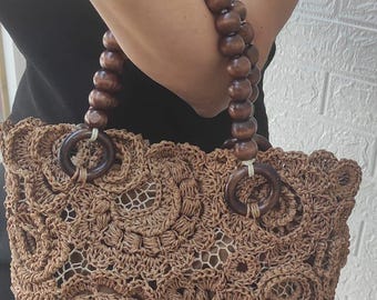 May include: A brown crochet tote bag with a floral design. The bag has wooden handles and wooden rings attaching the handles to the bag. The handles are made of large, round wooden beads.