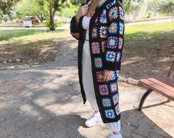 May include: A long, black crochet cardigan with a colorful patchwork design. The cardigan features a hood and is worn over a white top and pants. White sneakers complete the outfit. The background shows a park setting with trees and a paved walkway.